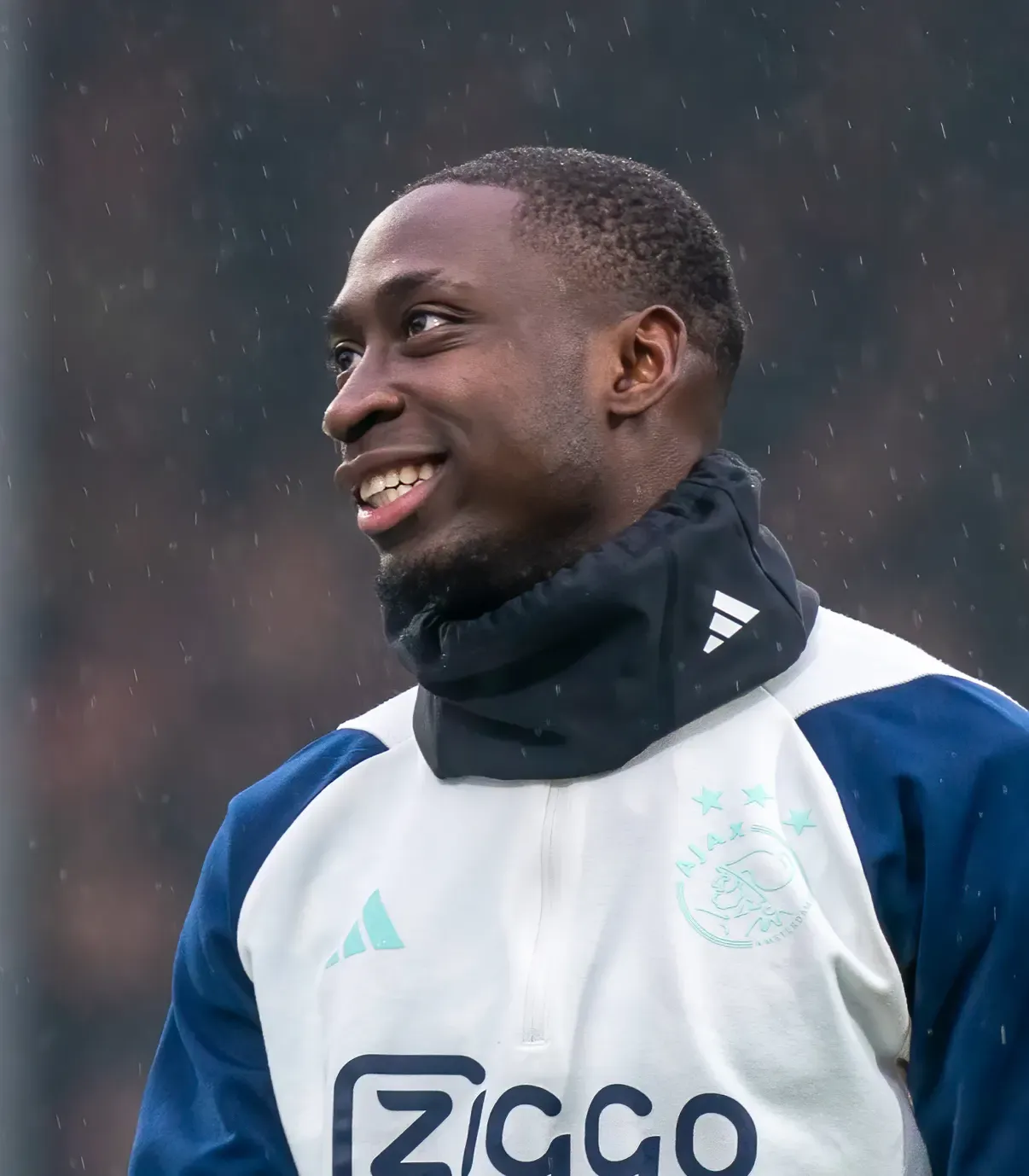 Carlos Forbs sorride a mezzo busto sotto la pioggia con scaldacollo e tuta dell'Ajax