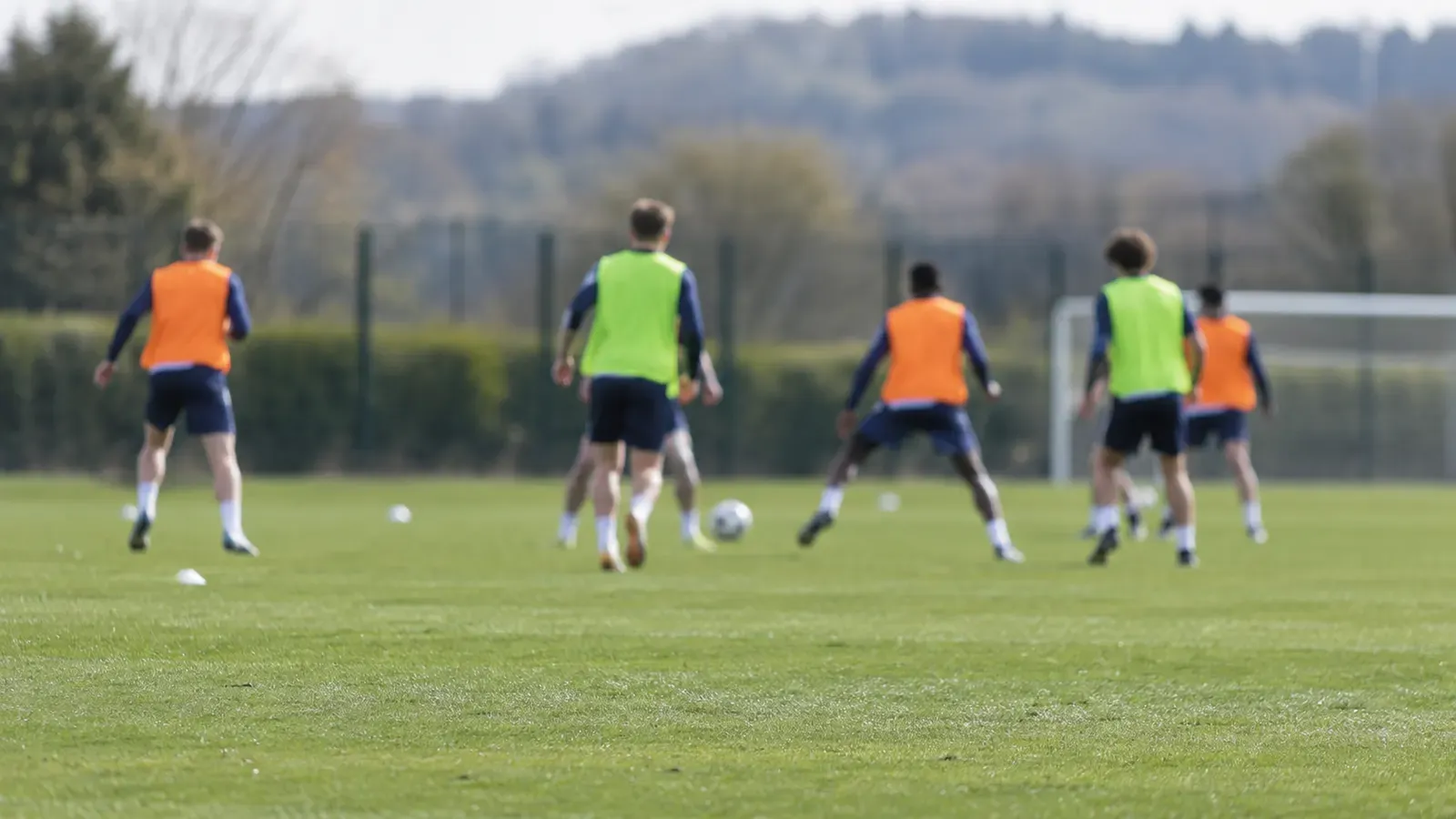 Foto generata con IA: Un campo di allenamento fotografato da bordo campo, con un piccolo gruppo di giovani calciatori lontani e sfocati durante un'esercitazione. I giocatori indossano abbigliamento sportivo senza scritte, numeri, loghi o sponsor; la luce è naturale e l'inquadratura ha il taglio di una fotografia giornalistica.