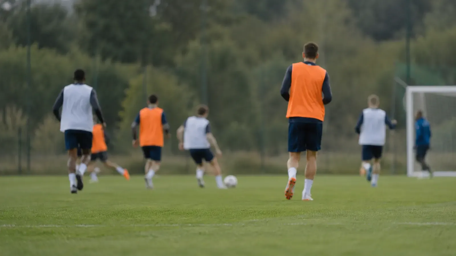 Foto generata con IA: Un campo di allenamento fotografato da bordo campo, con alcuni calciatori lontani e sfocati durante una seduta. Le figure sono riprese di spalle e indossano abbigliamento senza nomi, numeri, scritte o loghi, con luce naturale e taglio da fotografia sportiva.
