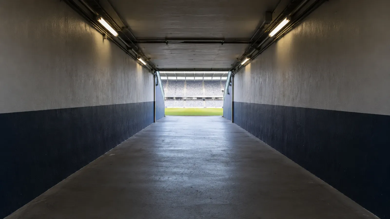 Foto generata con IA: Un tunnel degli spogliatoi vuoto, fotografato dal fondo verso l'apertura luminosa sul campo. Inquadratura grandangolare simmetrica, con luce artificiale sulle pareti e nessuna scritta, logo o elemento che identifichi un impianto preciso.