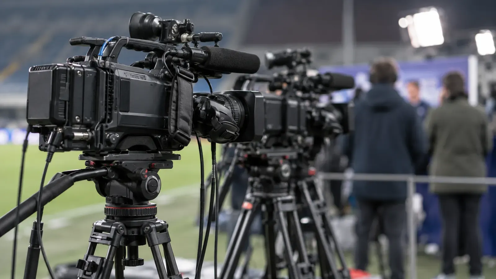 Foto generata con IA: Treppiedi e telecamere senza marchi in uno spazio predisposto per un’intervista sportiva, con operatori sfocati e di spalle sullo sfondo. Ripresa ravvicinata laterale, fuoco sull’attrezzatura e luce artificiale morbida, senza scritte, loghi o stemmi visibili.