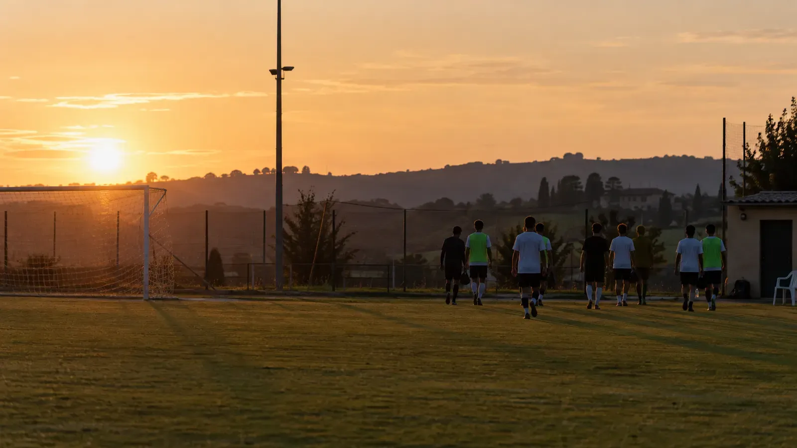 Foto generata con IA: Un gruppo di giovani calciatori visto da lontano e di spalle attraversa un piccolo campo al termine di una seduta, con le sagome in controluce. Ripresa laterale a campo lungo nella luce del tramonto, senza volti riconoscibili, scritte, numeri, marchi o stemmi.