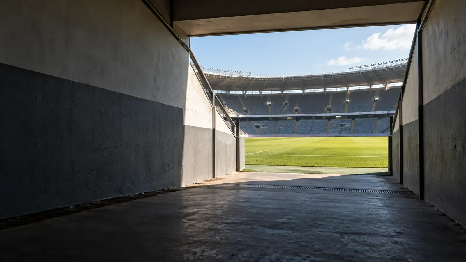 Foto generata con IA: Il tunnel vuoto di un impianto calcistico generico, con l’uscita verso il prato illuminata dalla luce del pomeriggio. Ripresa grandangolare dal basso e leggermente laterale, senza persone, scritte, loghi, stemmi o sponsor visibili.