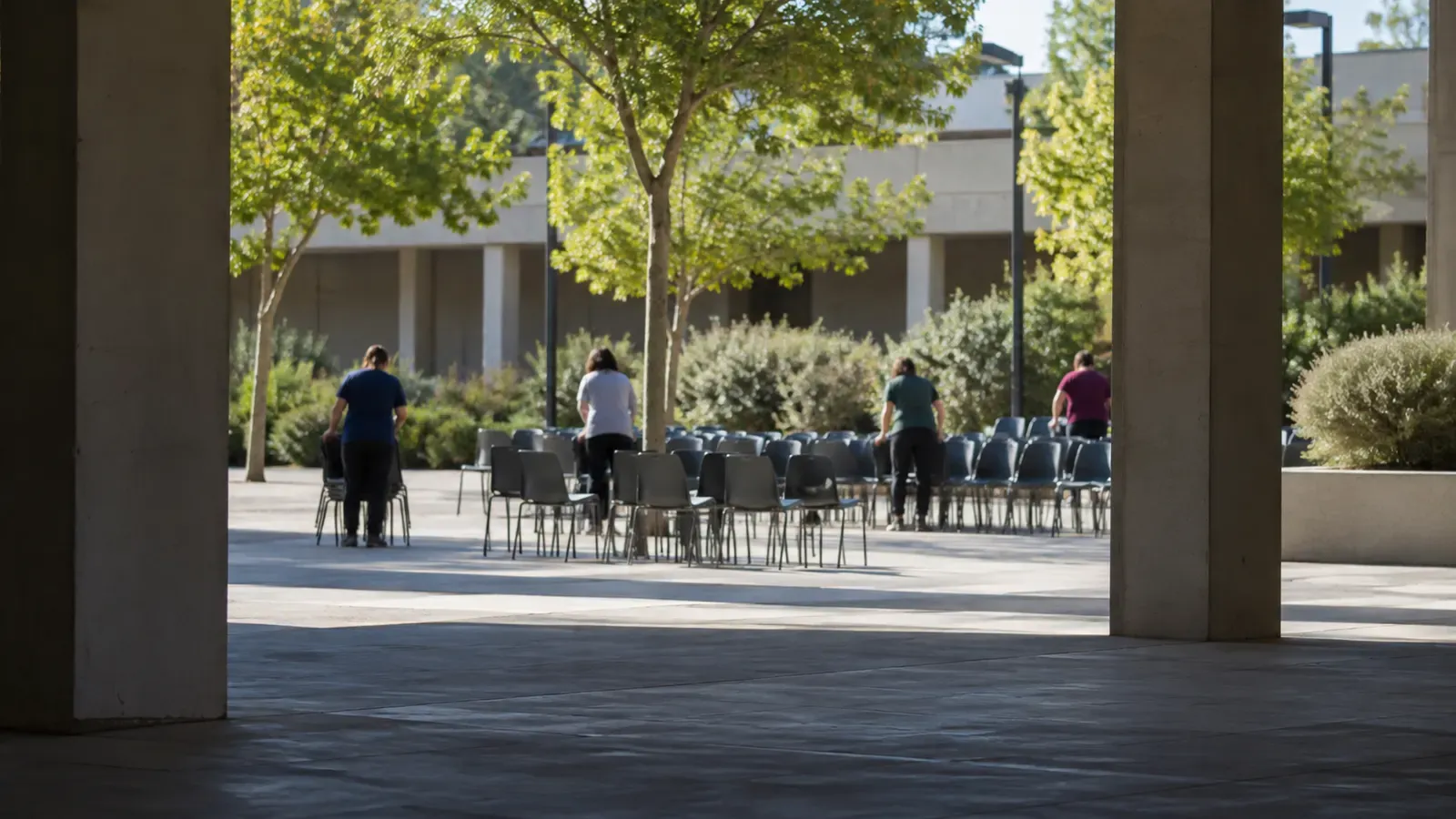 Foto generata con IA: Alcuni volontari visti di spalle sistemano sedie per l'accoglienza nel cortile di un centro sportivo generico. Ripresa obliqua da sotto un portico, nella luce naturale del mattino, con figure lontane e nessuna scritta, numero, marchio, stemma o sponsor visibile.