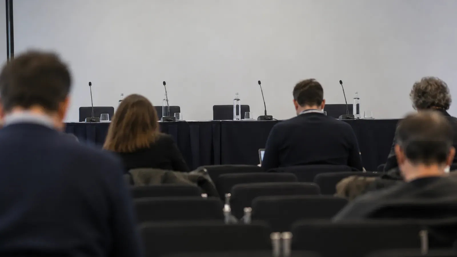 Foto generata con IA: Una sala stampa allestita per una presentazione, fotografata dal fondo con file di sedie e microfoni sul tavolo dei relatori. Alcune persone lontane e sfocate sono riprese di spalle, con luce naturale e senza scritte, loghi o immagini di progetti visibili.