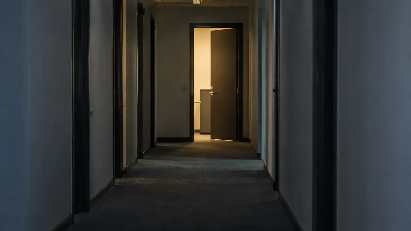 Foto generata con IA: Un corridoio di uffici con una porta socchiusa e luce accesa nella stanza in fondo, fotografato la sera con prospettiva centrale ad altezza d'occhio. Nessuna persona presente, nessuna scritta, insegna, logo o immagine di progetti visibile.