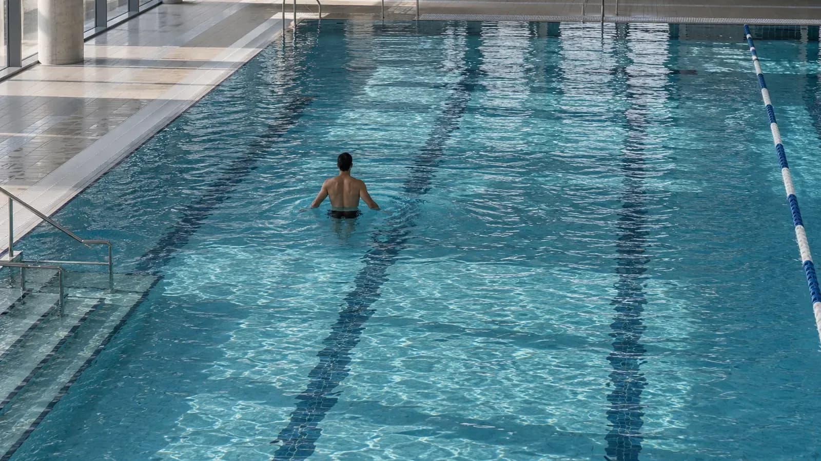 Foto generata con IA: Un atleta lontano e visto di spalle svolge un esercizio di recupero in una piscina di un centro sportivo. Ripresa dall’alto, con luce naturale riflessa sull’acqua, senza volti riconoscibili, scritte, marchi o stemmi.