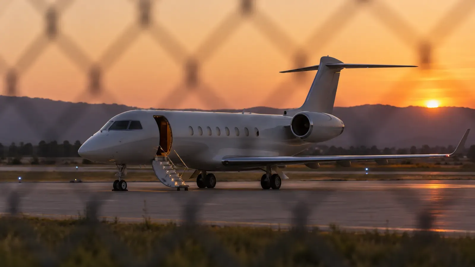 Foto generata con IA: Un jet fermo sulla pista di un aeroporto privato, con la scaletta vuota e nessuna persona visibile. Ripresa con teleobiettivo attraverso una recinzione al tramonto, senza scritte, marchi o loghi.