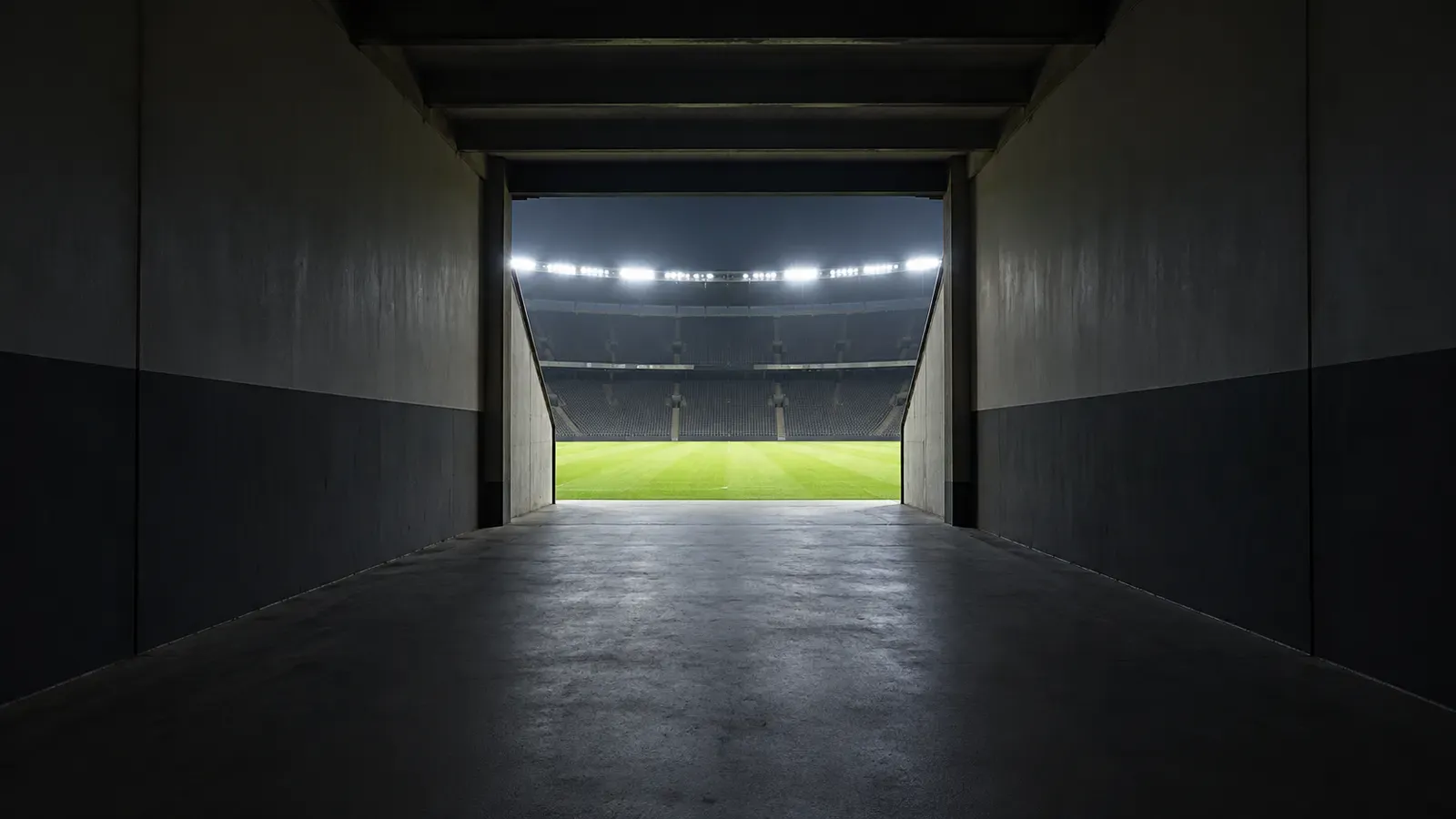 Foto generata con IA: Il tunnel degli spogliatoi di uno stadio visto dal fondo verso l'uscita sul campo, completamente deserto prima dell'incontro. Ripresa grandangolare simmetrica dalla penombra interna, con la luce intensa dei riflettori che illumina il rettangolo d'erba in lontananza. Nessuna persona, scritta, logo, stemma o sponsor presente nell'inquadratura.