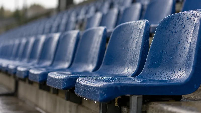 Foto generata con IA: Una fila di seggiolini bagnati sulle tribune di un impianto calcistico, con gocce ferme sui bordi e le file successive sfocate. Ripresa laterale con teleobiettivo nella luce dopo la pioggia, senza persone, scritte, numeri, loghi o elementi che identifichino uno stadio preciso.